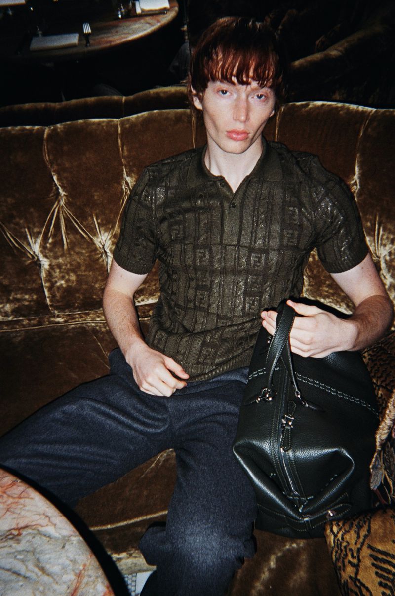 An adult male with auburn hair sits on a brown tufted couch holding a black handbag. The scene is dimly lit.