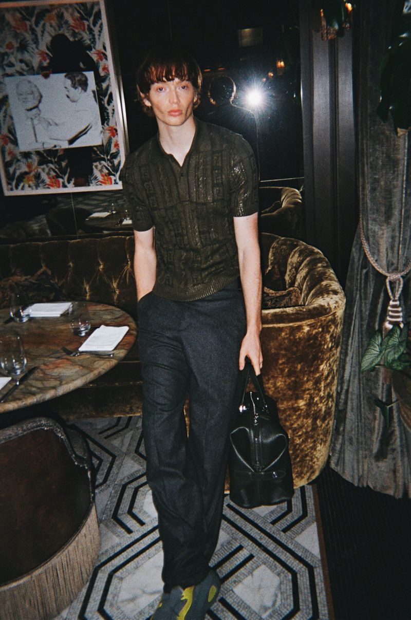 Man in patterned shirt and dark pants holding a bag in a warmly lit room with a patterned carpet and marble table.