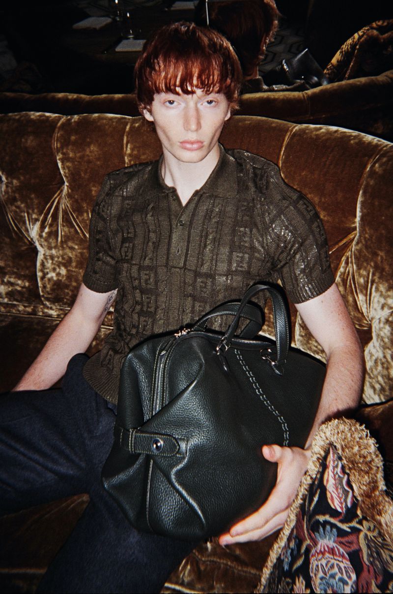 Male model sits on a velvet couch with a Fendi patterned shirt holding a black leather duffel bag in a studio setting. 