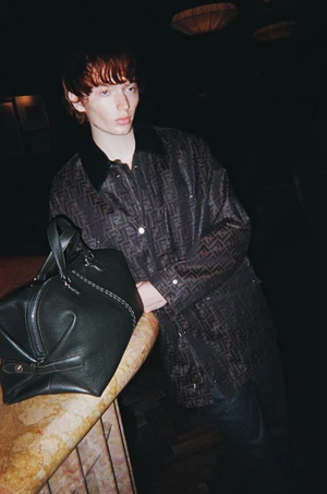 Model leaning against a bar counter next to a dark green leather handbag, wearing a monogram patterned jacket