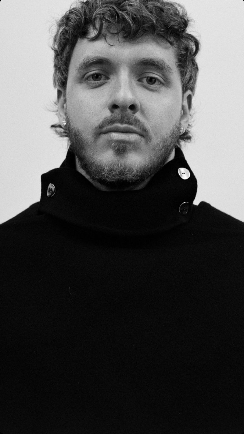 Black and white portrait of a man with curly hair wearing a black turtleneck sweater, looking directly at the camera with a neutral expression.