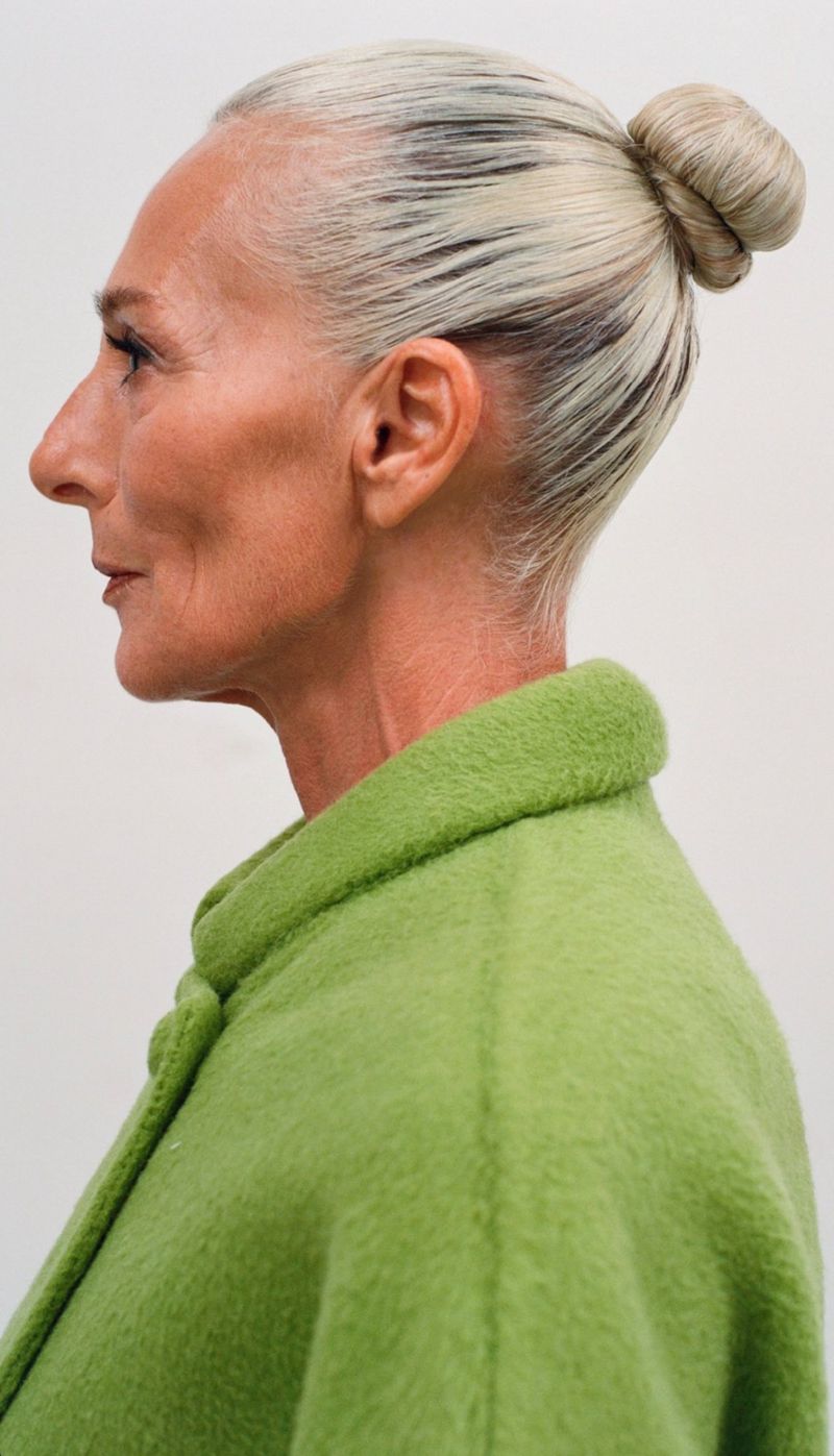 Profile shot of a senior woman with gray hair styled in a bun, wearing a light green coat against a white background in a studio setting.