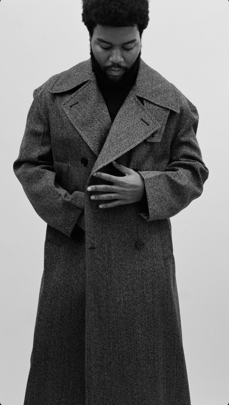 Monochrome portrait of a black male wearing a long overcoat and black turtleneck sweater, against a light gray background.