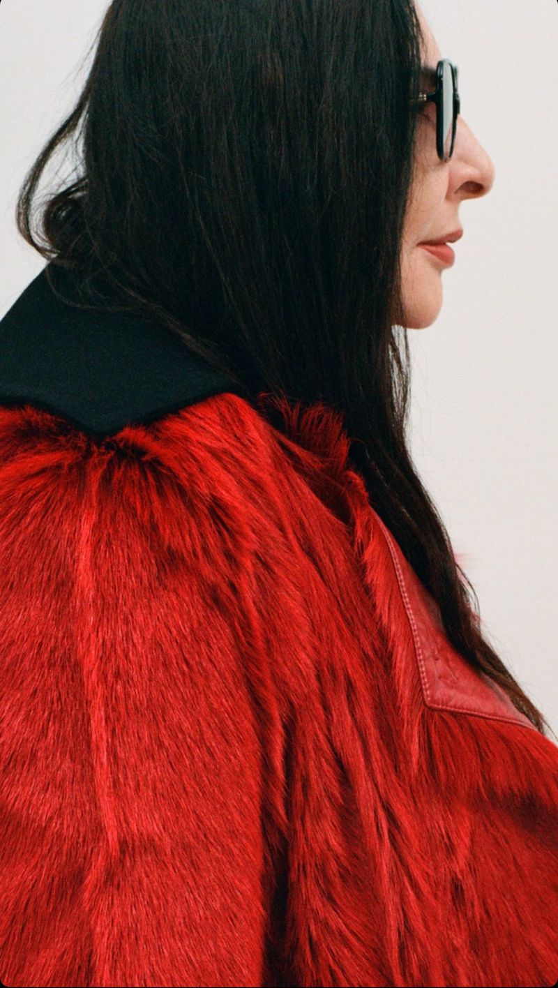 Profile shot of a woman with dark hair wearing a red fur coat and sunglasses against a neutral background in an indoor setting.