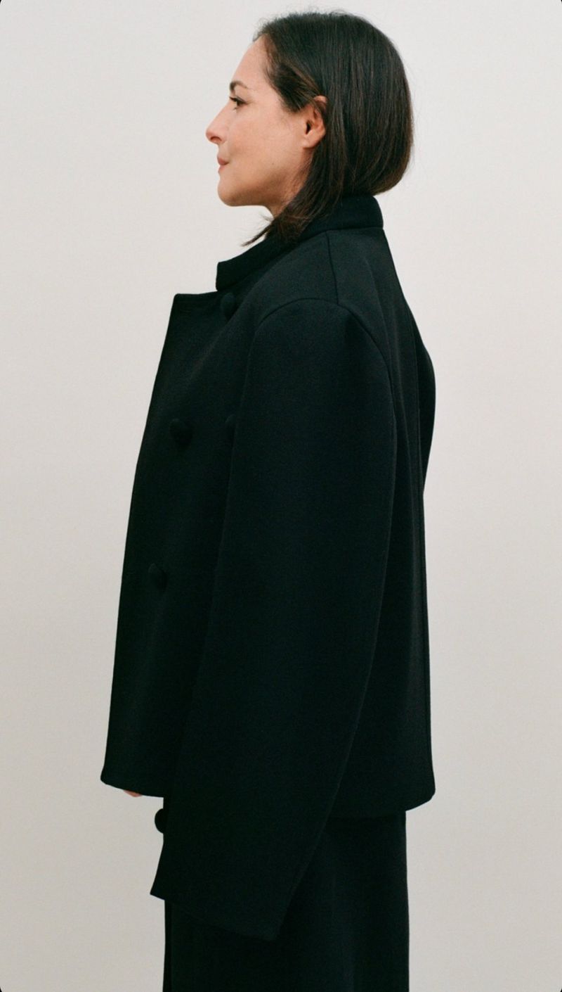 A woman in a black jacket is captured in a side profile shot against a neutral backdrop, highlighting fashion and simplicity.