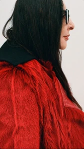 Profile portrait of a woman wearing black glasses and a textured red coat against a plain white background.