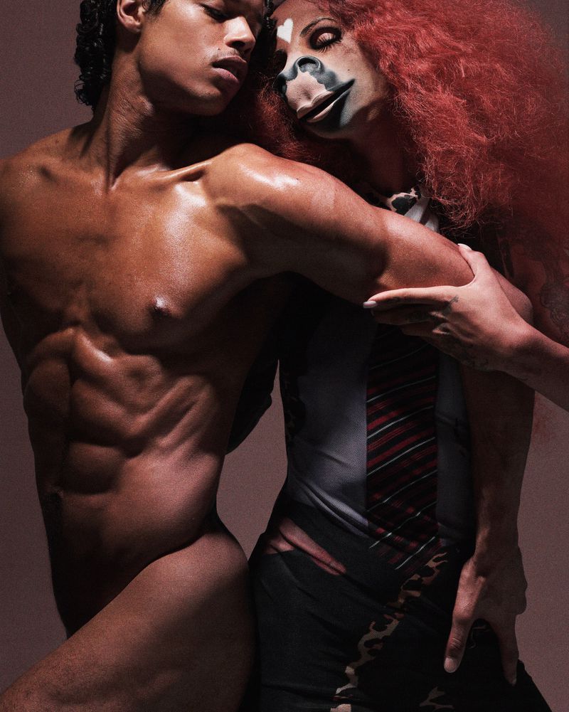 Striking fashion portrait of a muscular, shirtless man embracing a woman with theatrical makeup and red hair against a neutral backdrop.