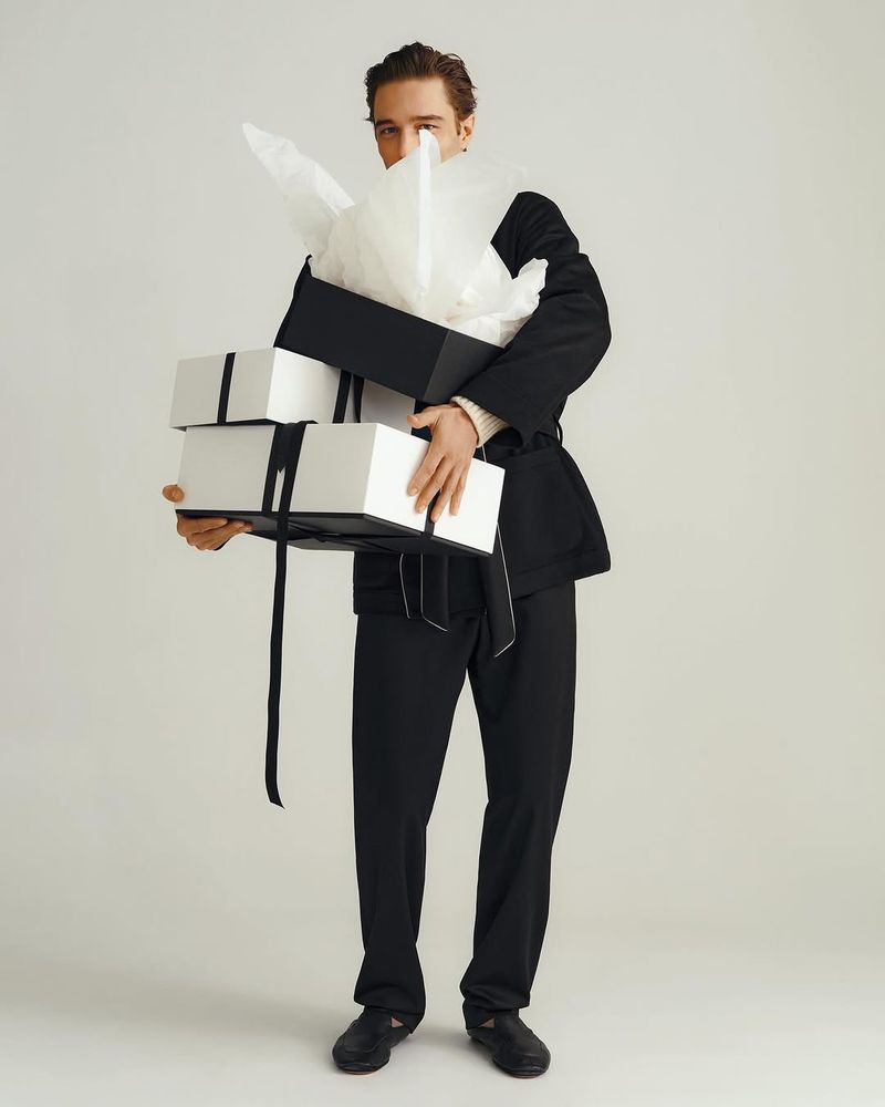 A man in a black suit holds a stack of black and white gift boxes in a brightly lit studio setting against a white backdrop.