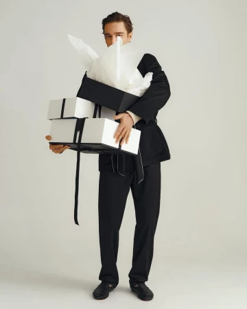 A man in a black suit holds a stack of black and white gift boxes in a brightly lit studio setting against a white backdrop.