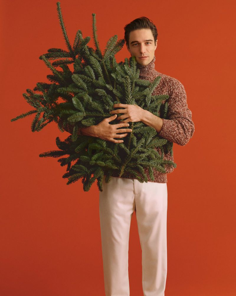 Young man in sweater embraces Christmas tree against orange backdrop, creating festive mood for holiday or seasonal fashion ad.