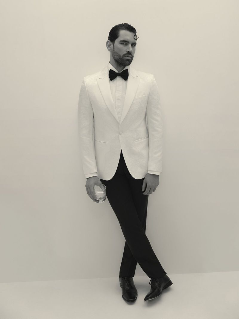 Monochrome portrait of a man in a white tuxedo with a bow tie holding a drink glass, standing in front of a neutral wall.