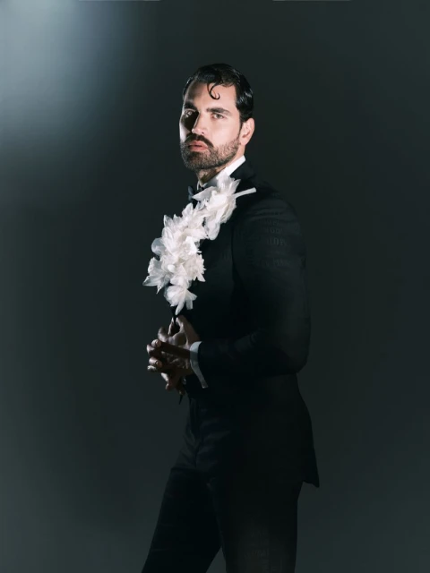 Portrait of a handsome adult male wearing a black tuxedo with a ruffled collar posing in a dimly lit photo studio setting.