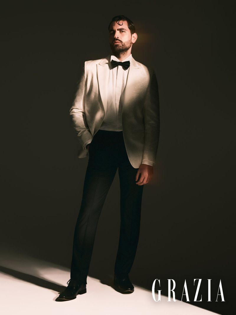 Portrait of an adult man in a white suit and black bow tie, standing in a photo studio with rim lighting. Wearing black dress shoes.