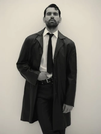 Model Emanuele Antonio Panico posing in a black leather trench coat, white collared shirt, and black necktie against a plain backdrop.