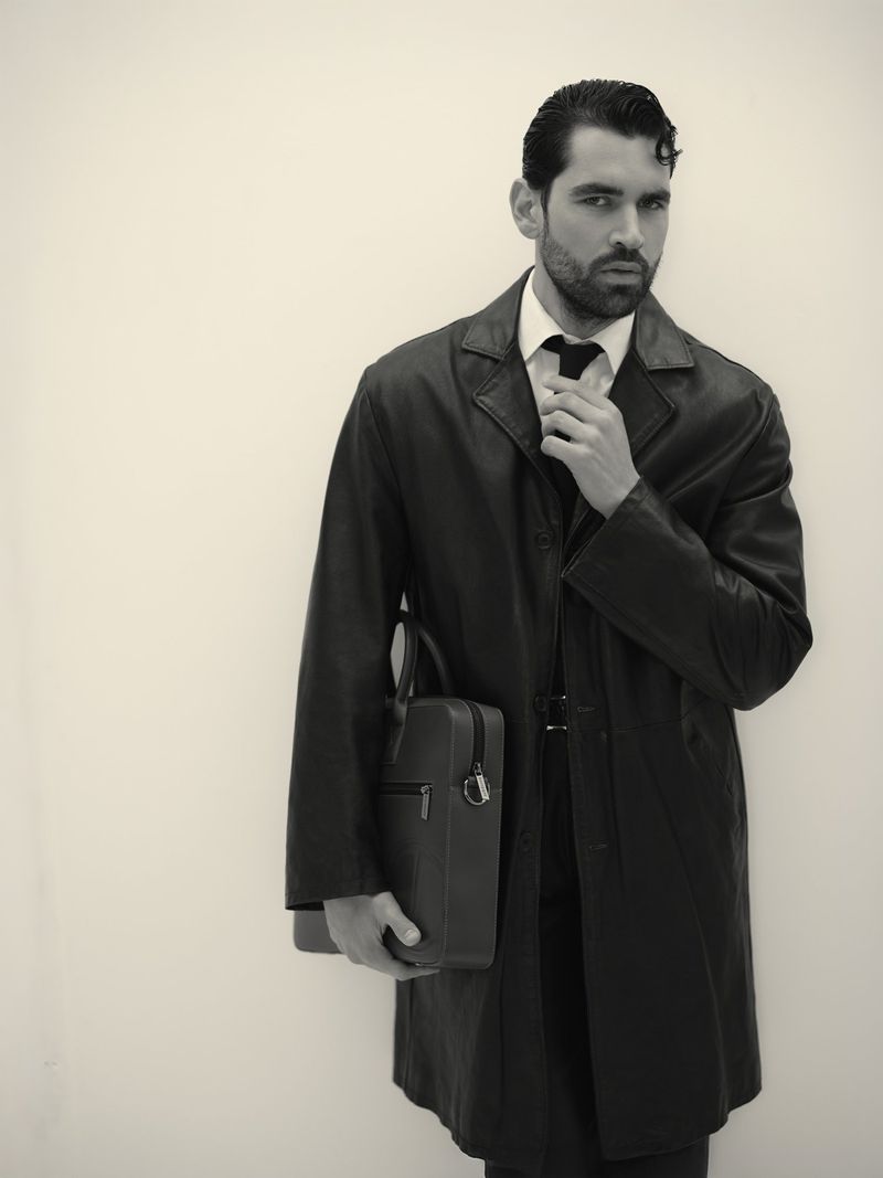 Monochrome portrait of an adult man in an overcoat, adjusting his tie, and holding a briefcase against a plain background.