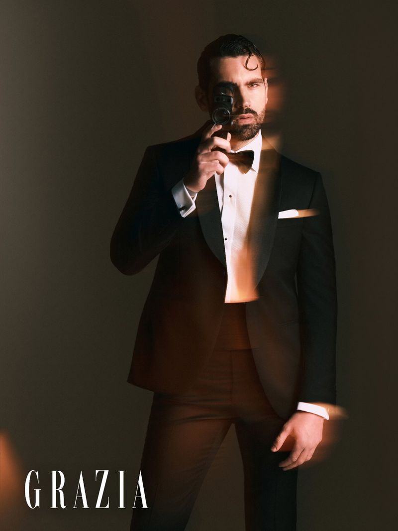 Stylish adult man in a black tuxedo and bow tie holds a vintage camera in a studio shot with Grazia magazine logo visible.