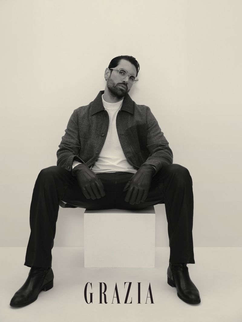 Monochrome portrait of an adult male in a tweed jacket and black trousers seated on a cube, posing with a serious expression.