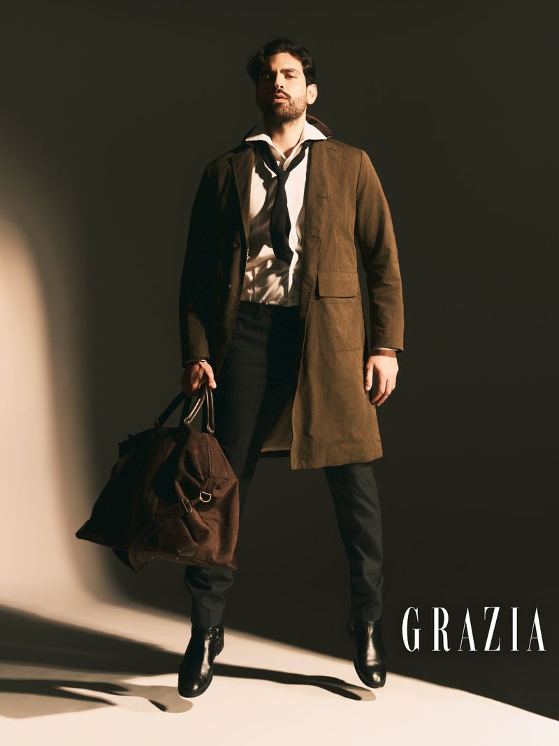 Model Emanuele Antonio Panico jumping mid-air in a brown overcoat holding a duffle bag against a shadowy studio backdrop for Grazia.