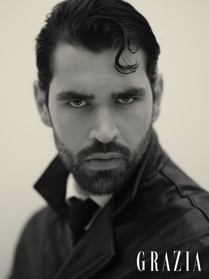 Black and white portrait of a man with dark hair and beard wearing a leather jacket against a plain background, with the Grazia logo.
