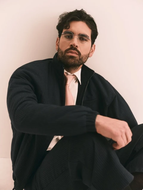Portrait of a dark-haired man with glasses, wearing a navy jacket, light pink shirt and tie, seated against a light peach background.