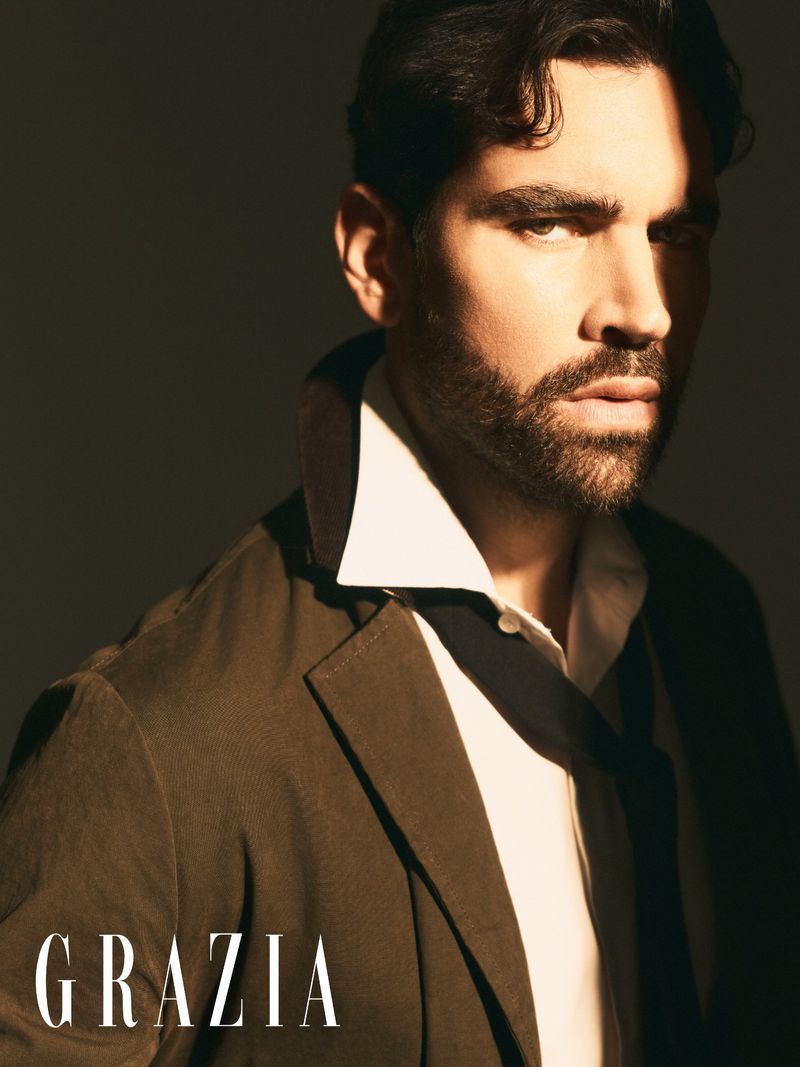 Portrait of model Emanuele Antonio Panico wearing an open brown jacket, popped white collar, and loosened dark tie in dramatic hard light.