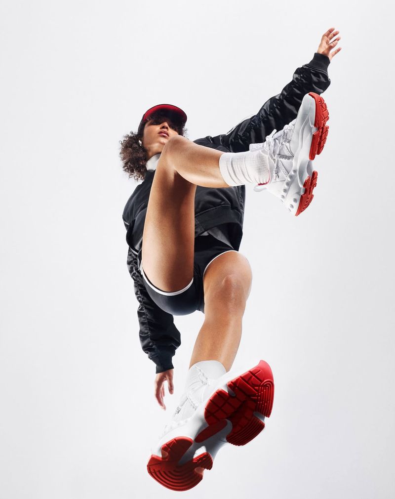 Athletic woman in black jacket, shorts, white socks, and red cap balancing in white and red sneakers against a clean white backdrop.