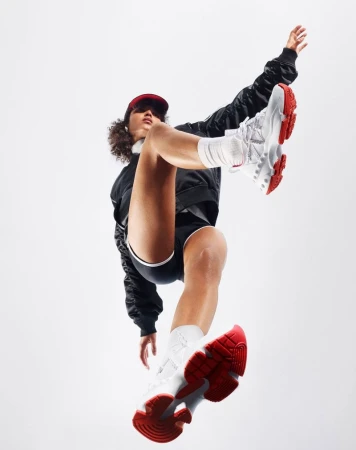 Athletic woman in black jacket, shorts, white socks, and red cap balancing in white and red sneakers against a clean white backdrop.