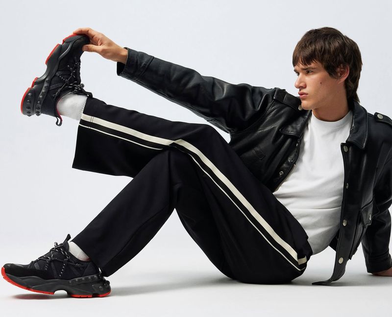 Stylish man poses while seated, wearing a black leather jacket, white t-shirt, black pants with white stripes, and black sneakers with red soles.