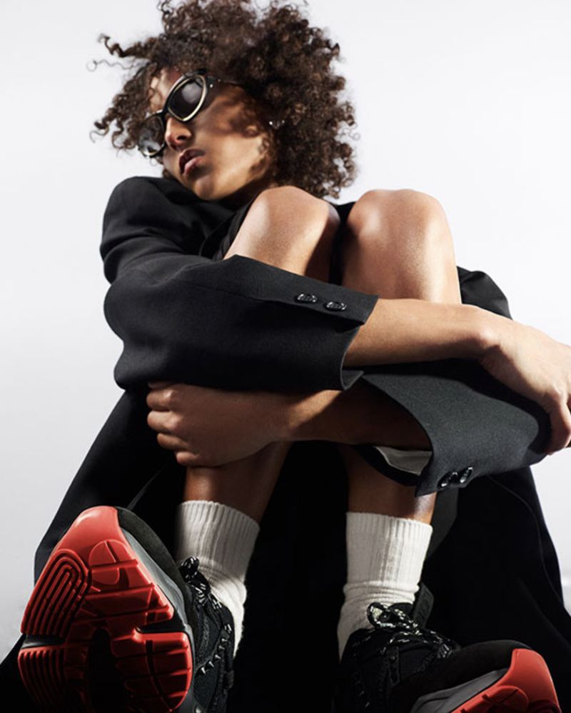 Fashionable young adult poses in a black blazer, socks, and trendy sneakers, set against a bright white studio backdrop.