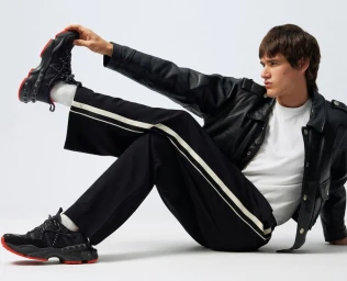 Male model wearing black leather jacket and striped trousers seated on white seamless backdrop stretching his sneaker