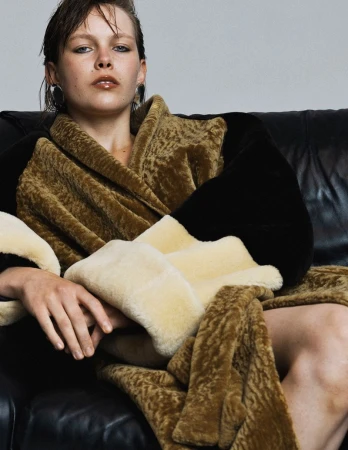 Model Thea Almqvist reclining in a textured mustard and black robe with cream faux fur cuffs on a black leather armchair.