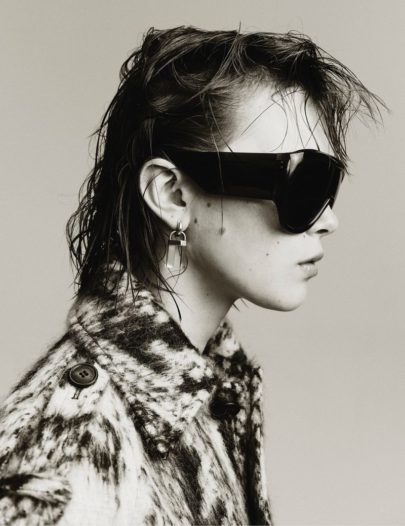 Black and white profile portrait of a woman with sunglasses, short hair, and an patterned coat against a neutral backdrop.