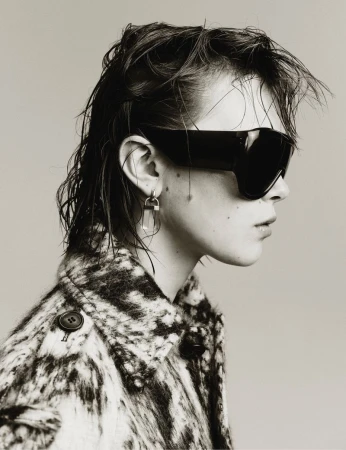 Black and white profile portrait of a woman with sunglasses, short hair, and an patterned coat against a neutral backdrop.