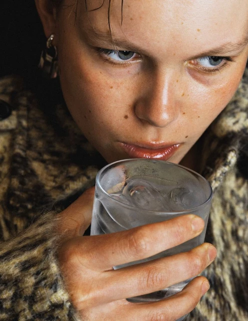Portrait of a young adult with freckles, drinking water from a textured glass, wearing a patterned jacket indoors in studio lighting.