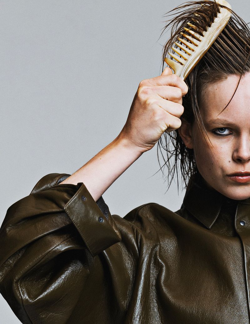 Close-up shot of a fair-skinned adult woman in a green leather jacket, styling her wet, short hair with a beige comb against a grey backdrop.