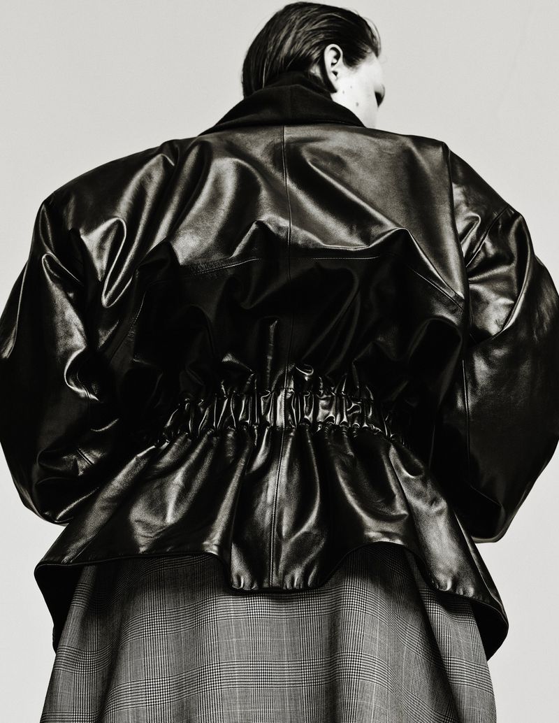 Back view of a person wearing a glossy black leather jacket with a gathered waist and a gray plaid skirt, against a neutral background.