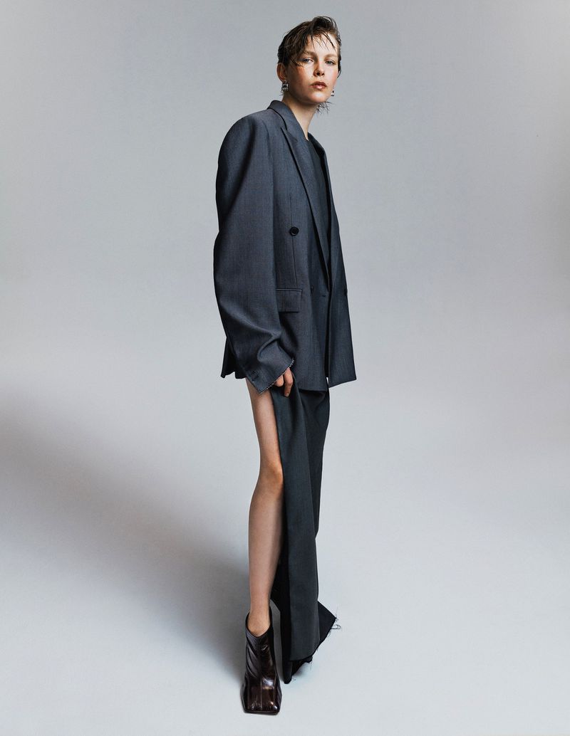 Fashion shot of a woman with short hair wearing an oversized grey suit with dark brown boots, posing in a full shot in a studio.