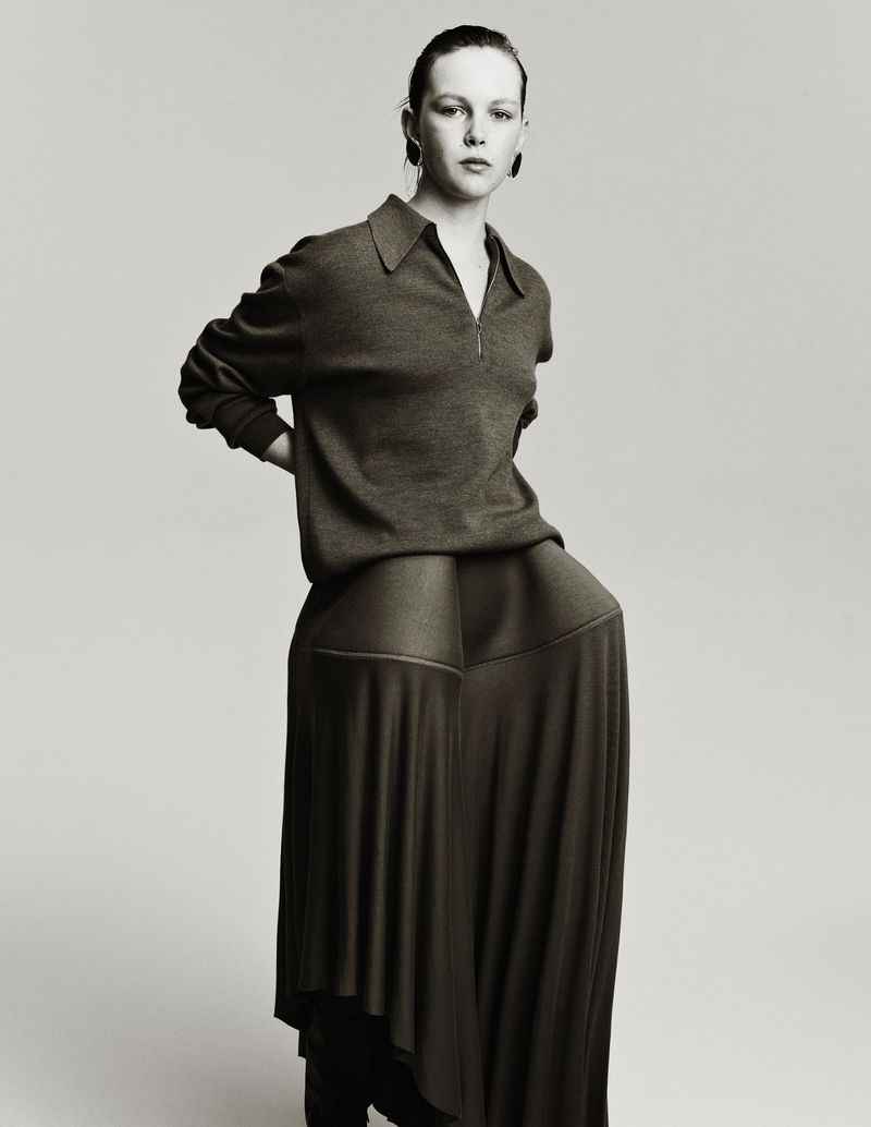 Monochrome portrait of a young woman in a long-sleeved top with a zipper, paired with a matching skirt, posing in front of a gray backdrop.
