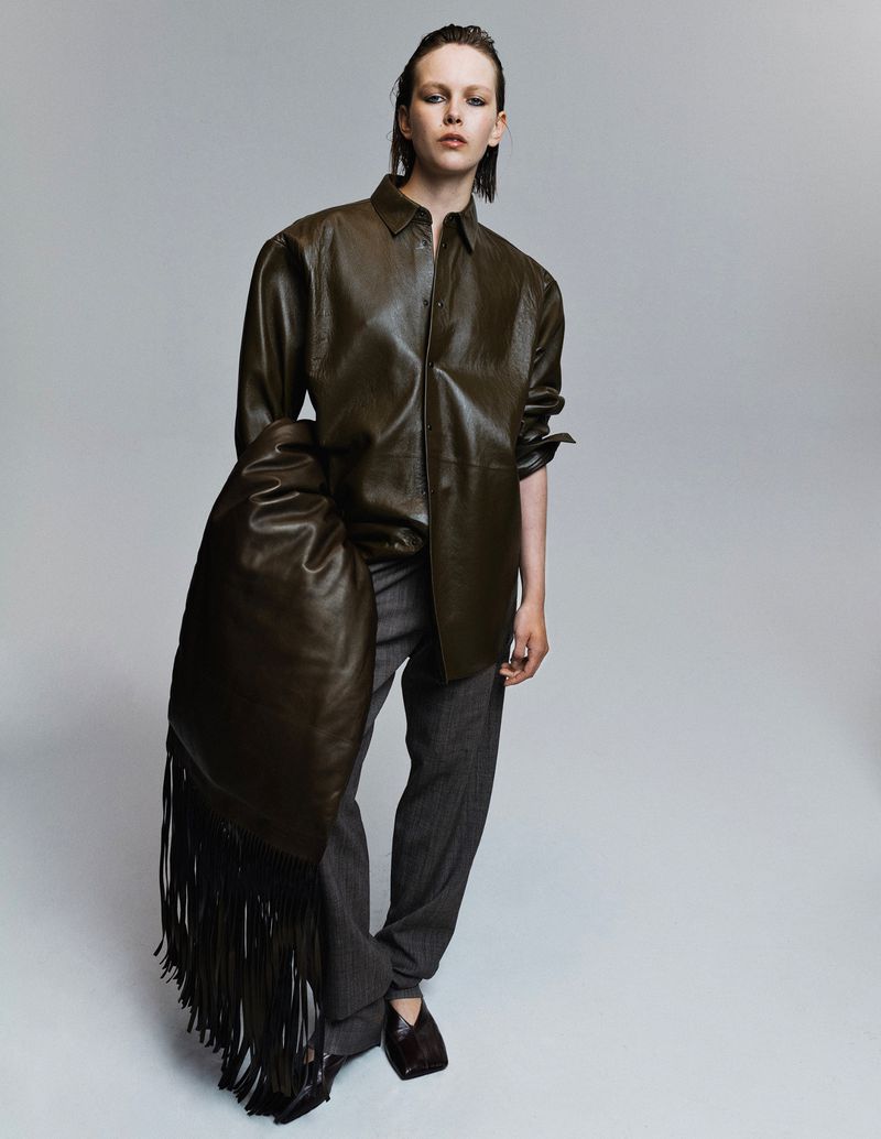 Fashion shot of a model in a green leather jacket, gray pants, and dark shoes posing with a large fringe leather bag in a studio.