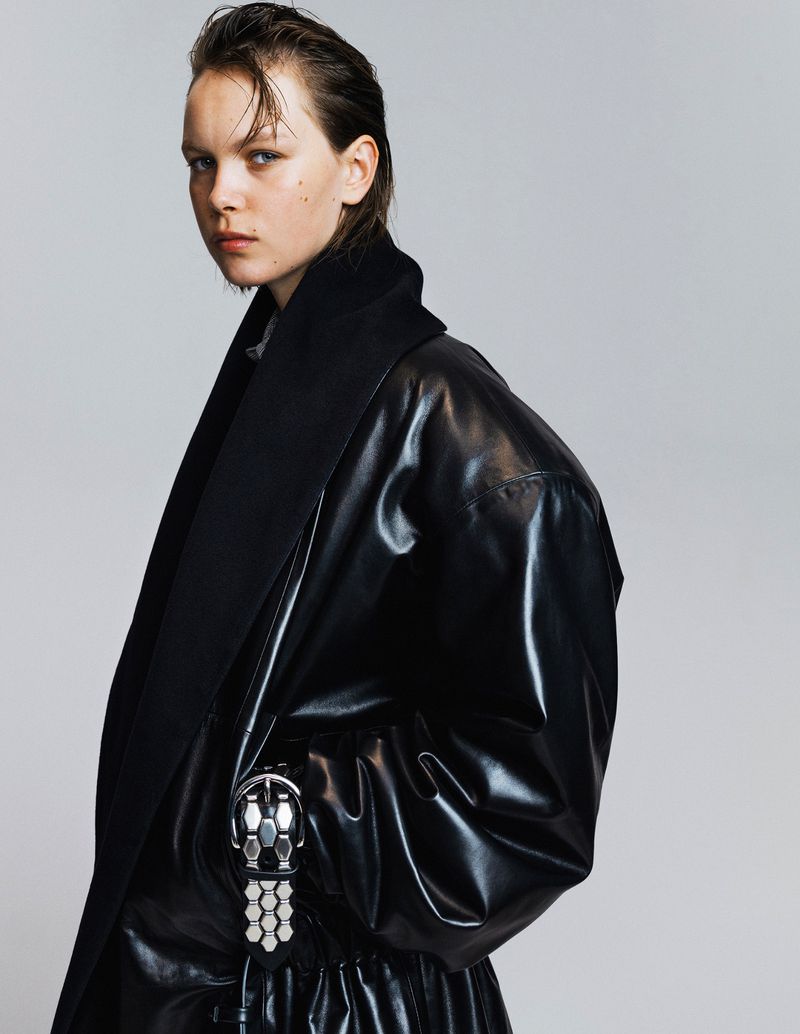 Woman in a black leather-look coat with a large decorative buckle, striking a pose against a plain light-grey background in a studio shot.