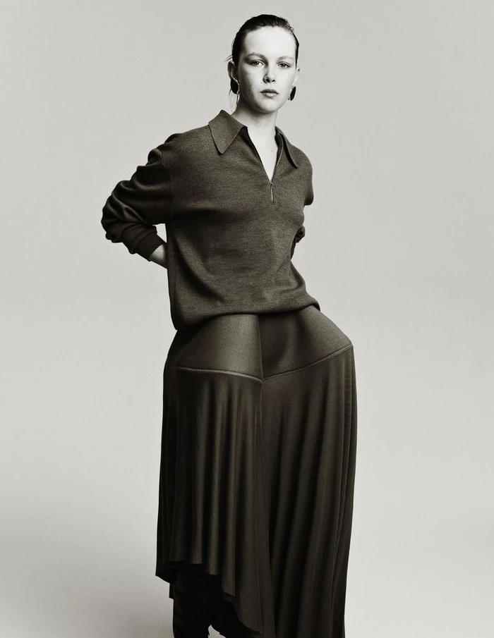 Model Thea Almqvist wearing a collared polo sweater and sculptural pleated wide-leg trousers against a neutral studio backdrop.