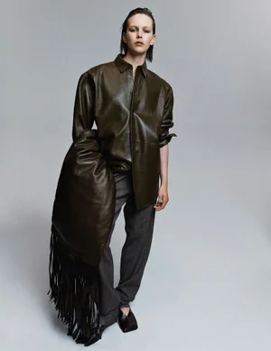 Model Thea Almqvist wearing an oversized dark olive leather shirt and grey trousers with a fringe accessory against a grey background.