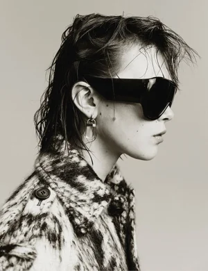 Profile of a woman with wet hair wearing oversized black shield sunglasses and a patterned black and white coat against a studio backdrop.