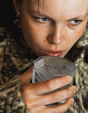 Close-up portrait of model Thea Almqvist holding a glass of iced water near her lips while wearing a textured knit garment.