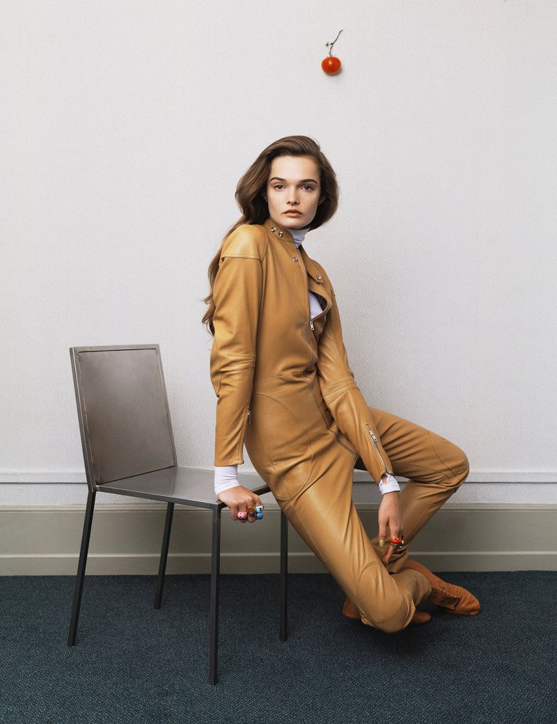 A woman in a tan leather suit poses next to a metal chair against a white wall, with a tomato attached to the wall above.