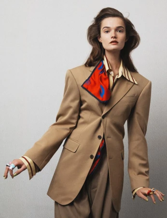 Portrait of a fashionable woman in a tan blazer and trousers, accessorized with colorful rings and a red scarf against a neutral backdrop.