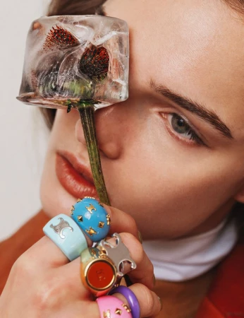 Close-up of a woman with fair skin and light eyes holding a flower encased in ice against her eye and wearing many colorful rings.