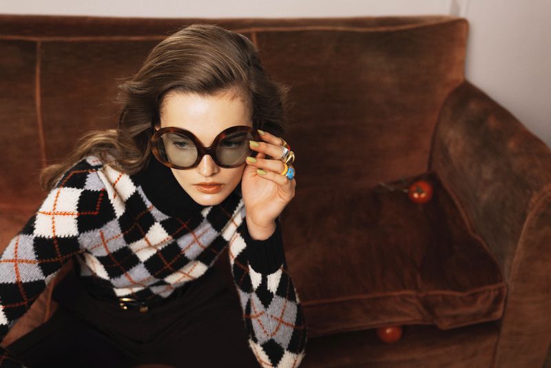Woman wearing glasses and argyle sweater sitting on a brown velvet couch with tomato props in a dimly lit indoor setting.