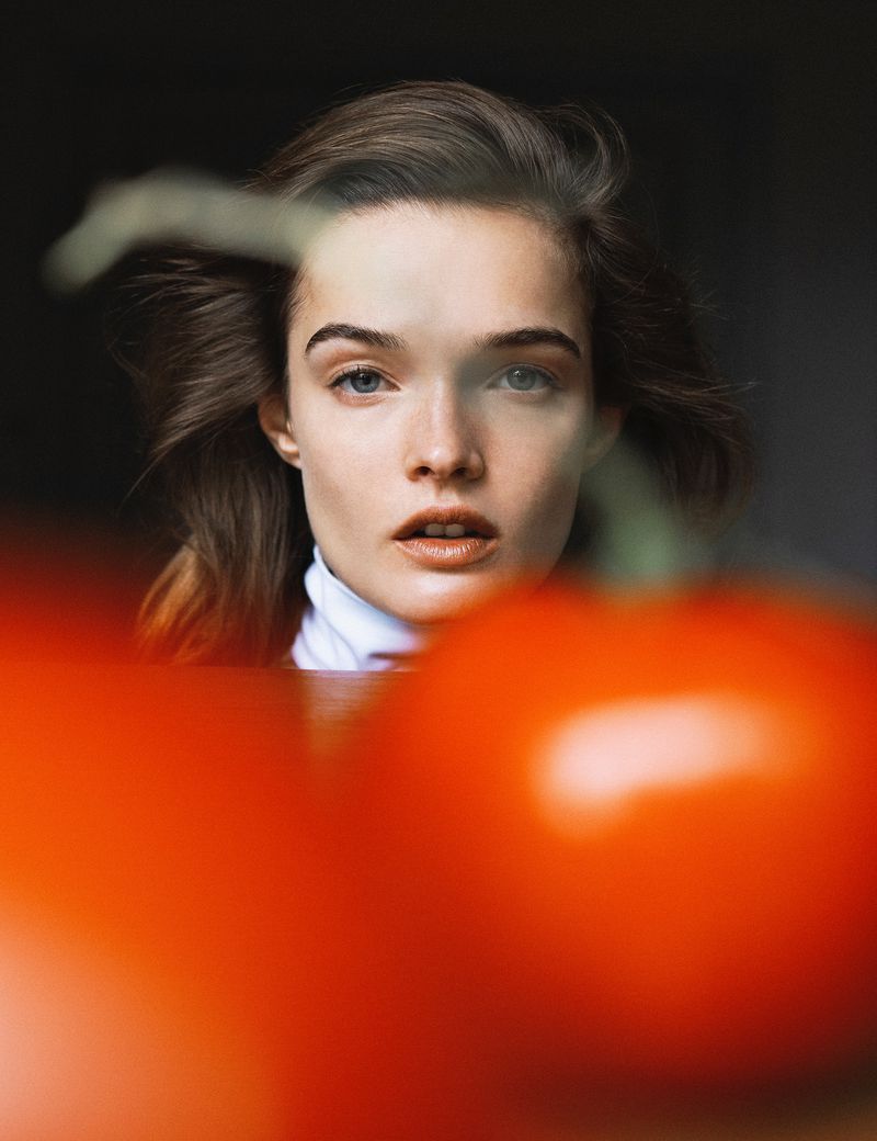 Portrait of a brunette woman with fair skin looking at the camera, framed by a blurred, out-of-focus tomato in the foreground.