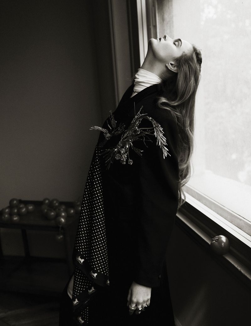Monochrome portrait of a woman in a dark coat standing by a window with decorative accents. Interior setting with natural light.
